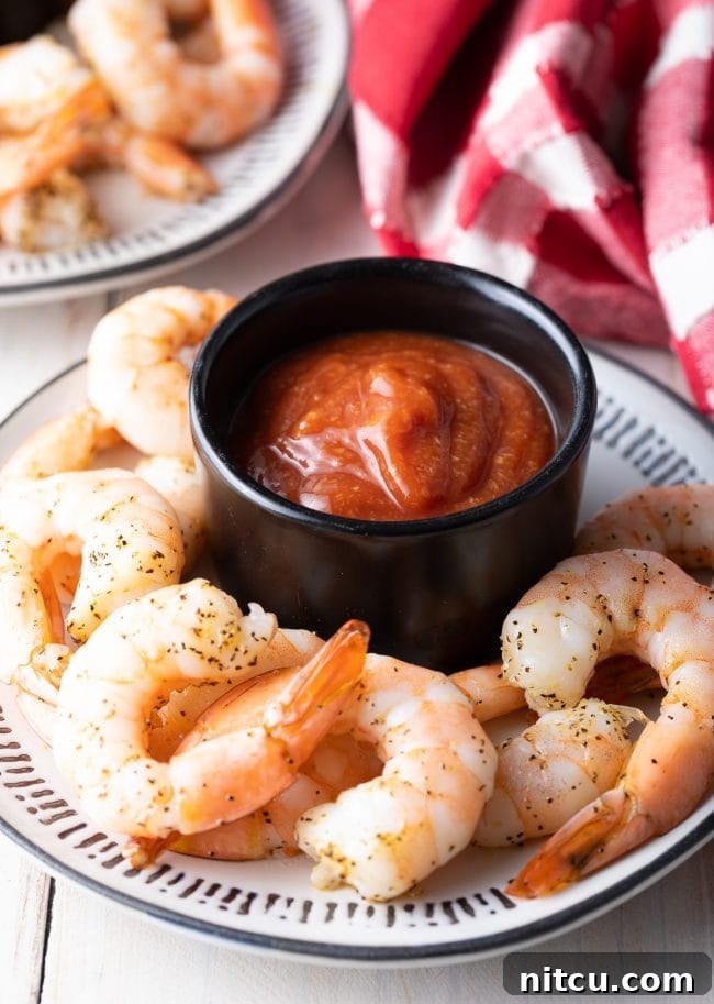 The best homemade cocktail sauce served in an elegant black ramekin, surrounded by perfectly cooked shrimp on a pristine white plate.