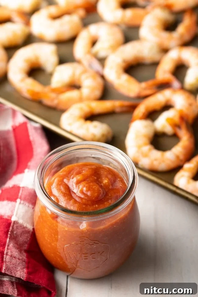 Homemade cocktail sauce glistening in a glass container, with a sheet pan of roasted shrimp blurred in the background, hinting at delicious possibilities.