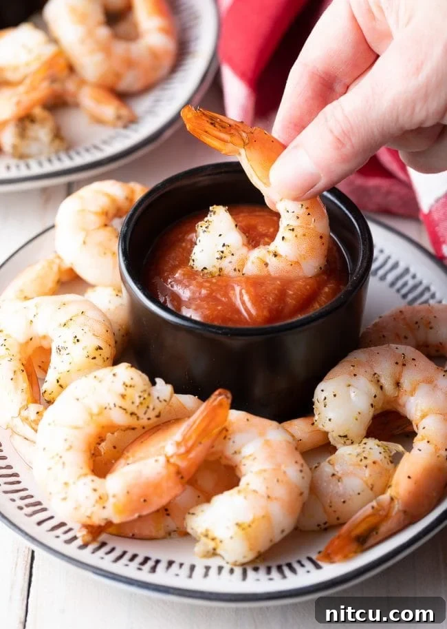 A hand gracefully dipping a large, cooked shrimp into a small bowl of homemade cocktail sauce, highlighting the perfect pairing.