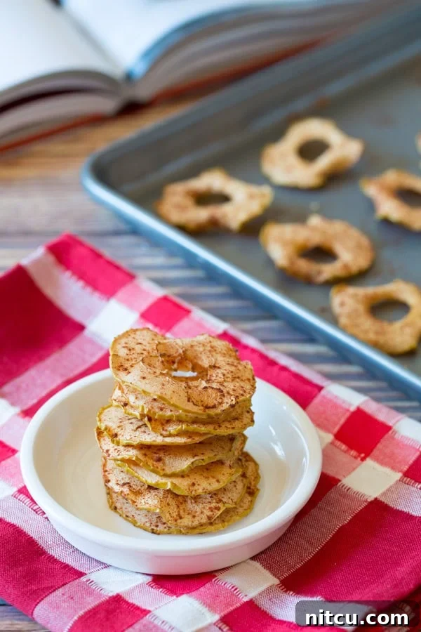 Golden Apple Crisps 3 Baked Apple Chips stacked up in a white bowl sitting on a red and white linen. More chips are in the background on a baking sheet.