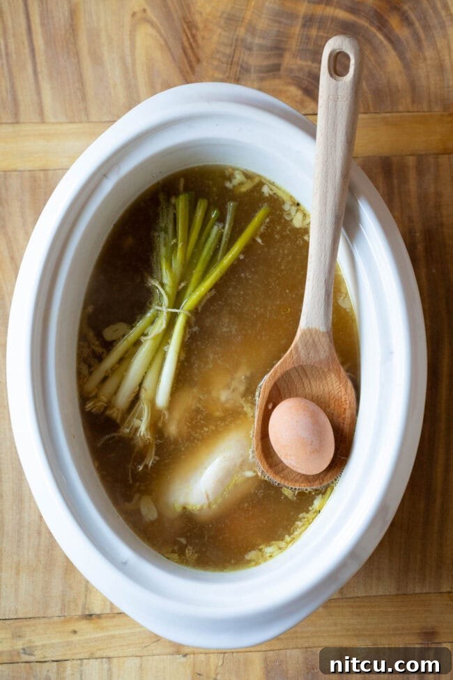 Raw ingredients, including chicken breasts, whole eggs, ginger, garlic, and scallions, being placed into a slow cooker pot, ready for the initial cooking stage of the ramen.