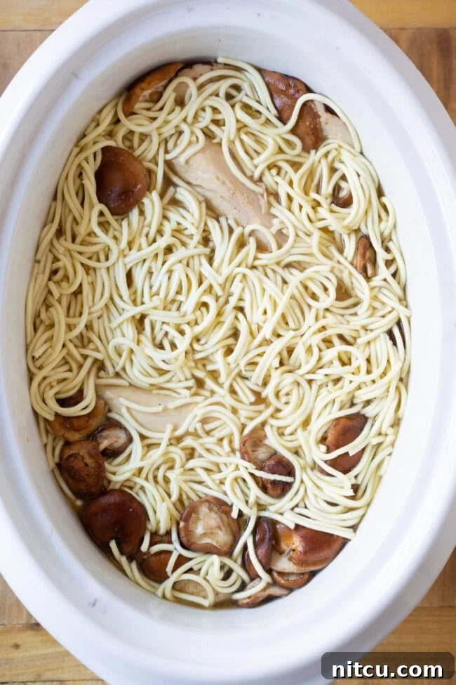An overhead shot of dried ramen noodles and shiitake mushrooms added into the slow cooker, with cooked chicken breasts placed on top to ensure noodles are submerged in the rich broth.