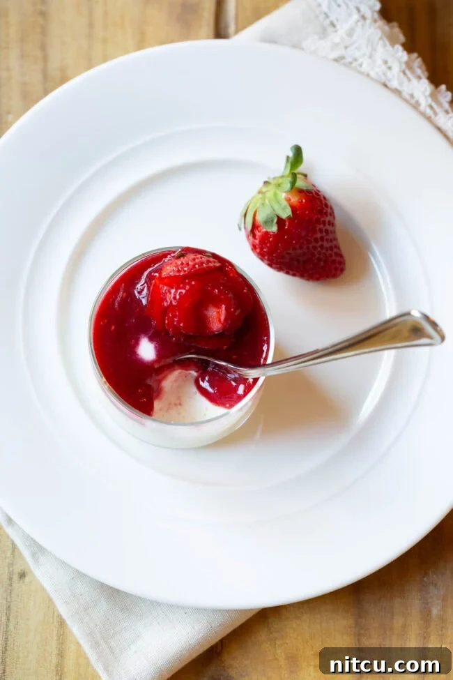 Top down view of a small glass dish with silky Swedish Creme, berry compote, and a strawberry rose. A metal spoon is elegantly scooping a portion from the dish.