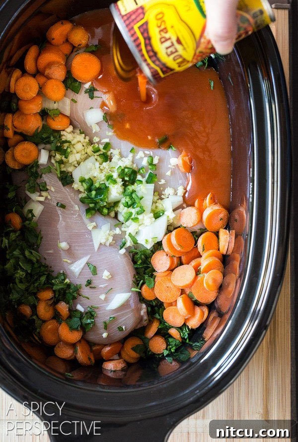 An overhead view of a slow cooker filled with raw chicken breasts, thinly sliced carrots, chopped onion, minced garlic, fresh cilantro, and a hand pouring Old El Paso Red Enchilada Sauce from a can over the ingredients, ready for cooking.