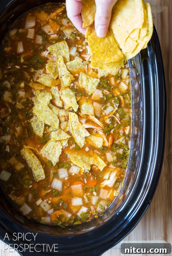 A hand gently adding crushed corn tortilla chips into a slow cooker filled with steaming chicken tortilla soup, ready to be stirred in for texture and flavor.