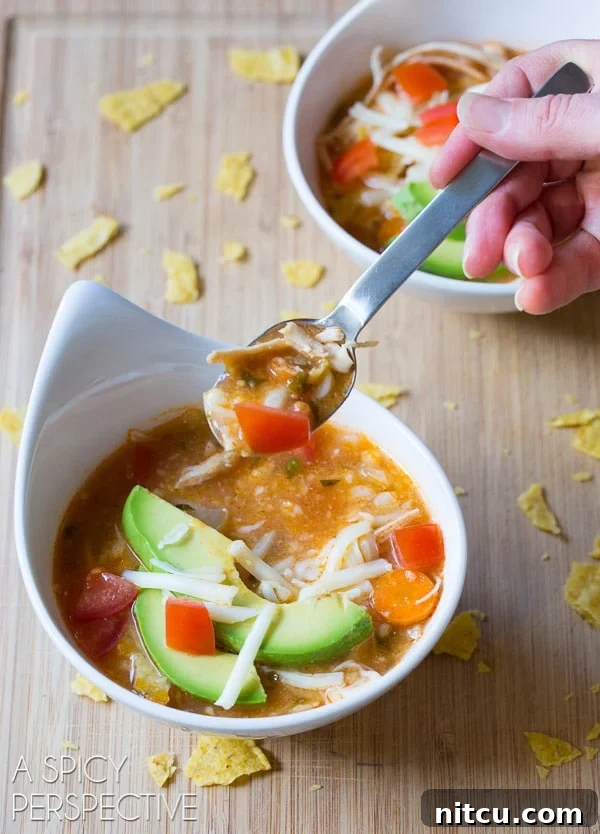 A hand holding a spoon, scooping a delicious bite of Slow Cooker Chicken Tortilla Soup from a white bowl, showing the rich broth, shredded chicken, and fresh toppings.