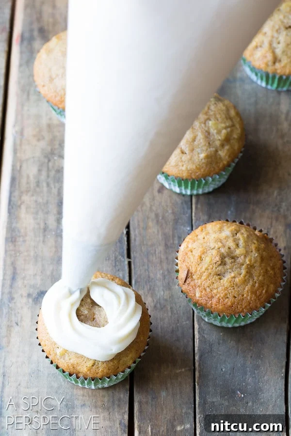 Luscious Hummingbird Cupcakes 6 A hand using a piping bag to swirl creamy sour cream frosting onto a baked Hummingbird cupcake.