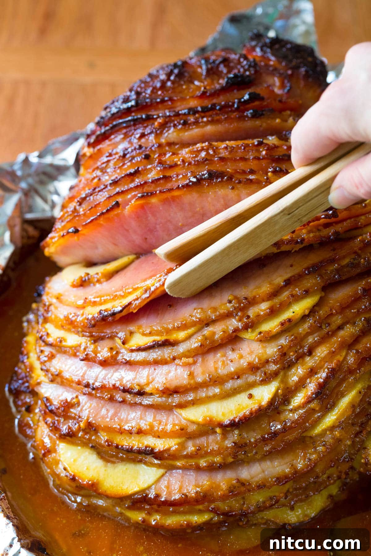 Honey-Glazed Ham with Roasted Apples 5 Close-up of tongs delicately removing a perfectly glazed and apple-stuffed slice of ham from the baking dish, highlighting its tender texture.