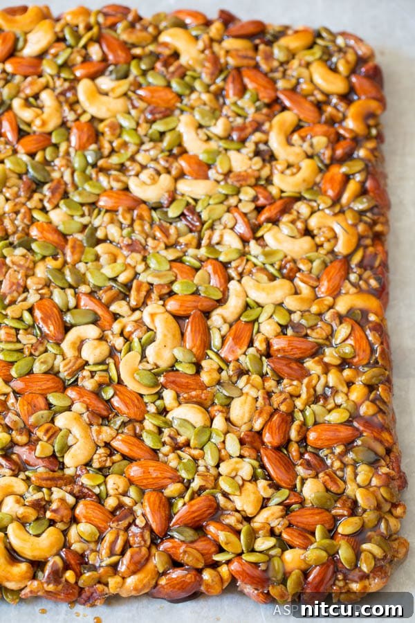 A large, golden-brown slab of baked nut bars, generously studded with pepitas, showcasing a rich, satisfying texture before cutting.