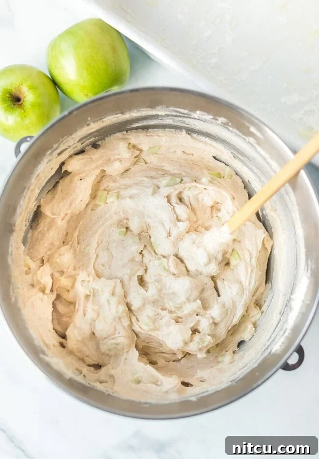 A spatula gently mixing diced apples into the prepared cake batter in a large mixing bowl.