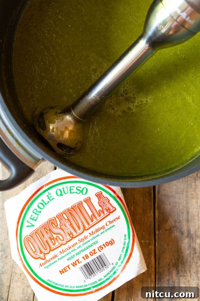 Immersion blender making the green soup creamy in a large pot. Block of quesadilla cheese is next to the pot.
