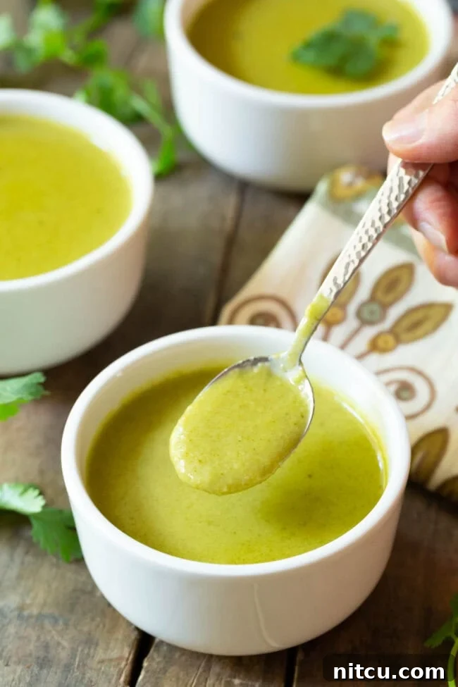 Hand with a metal spoonful of creamy green soup over a small white bowl, showing to camera.