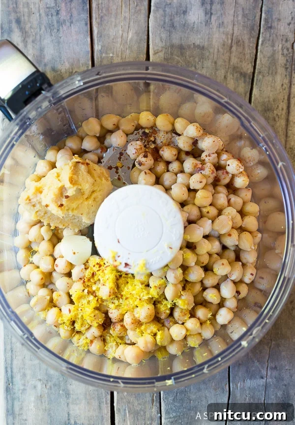 Umami Miso Hummus 3 Top down view food processor with chickpeas, lemon zest, scoop of miso paste, and cloves of garlic.