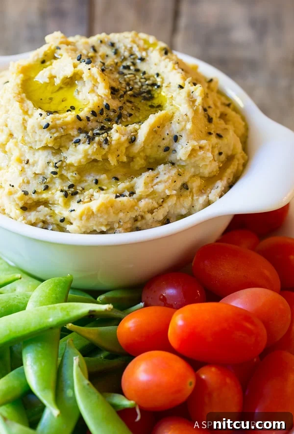 Umami Miso Hummus 5 Close view of a white bowl overflowing with homemade miso paste hummus garnished with sesame seeds. Around the dish are fresh sweet peas and grape tomatoes.