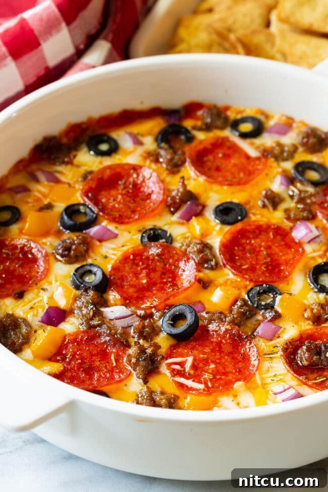 Fully Loaded Pizza Dip 2 White round dish with loaded supreme pizza dip, topped with pepperoni slices, ground cooked sausage, black olive slices, and chopped red onion.