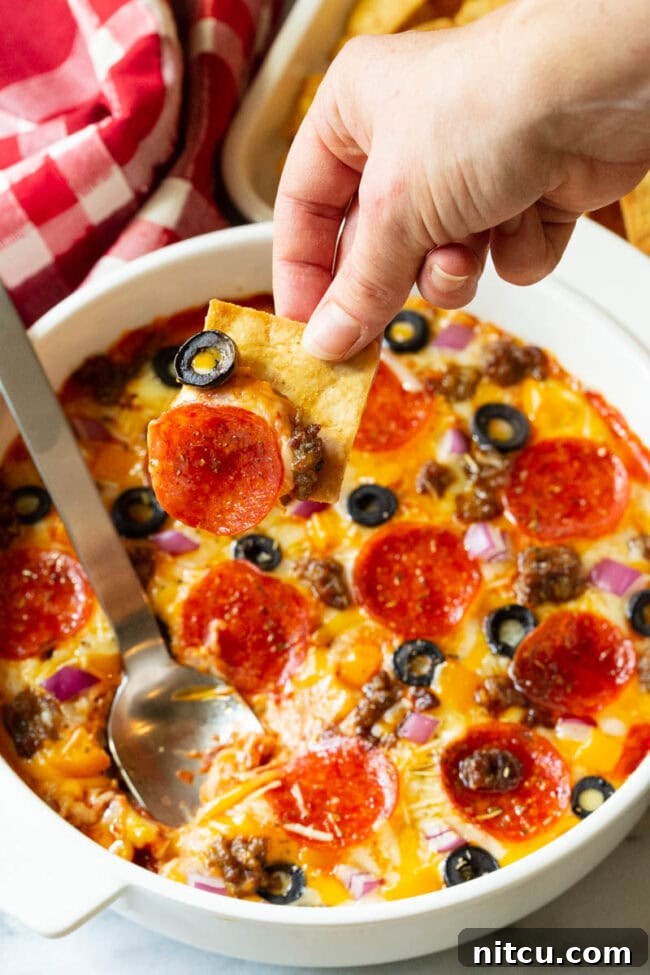 Fully Loaded Pizza Dip 3 White round dish with loaded supreme pizza dip, topped with pepperoni slices, ground cooked sausage, black olive slices, and chopped red onion. A hand is scooping a bite with a square pita cracker.