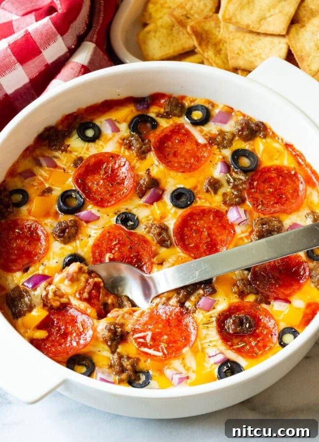 Fully Loaded Pizza Dip 4 White round dish with loaded supreme pizza dip, topped with pepperoni slices, ground cooked sausage, black olive slices, and chopped red onion.