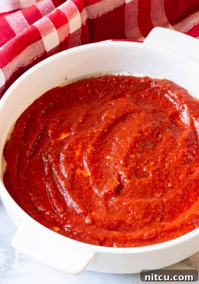 Fully Loaded Pizza Dip 6 Tomato sauce spread on top of cheese in a white round baking dish.