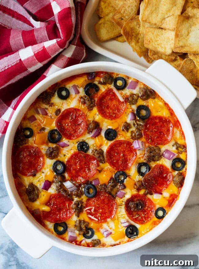 Fully Loaded Pizza Dip 8 Top down view of a white round dish with loaded supreme pizza dip, topped with pepperoni slices, ground cooked sausage, black olive slices, and chopped red onion.