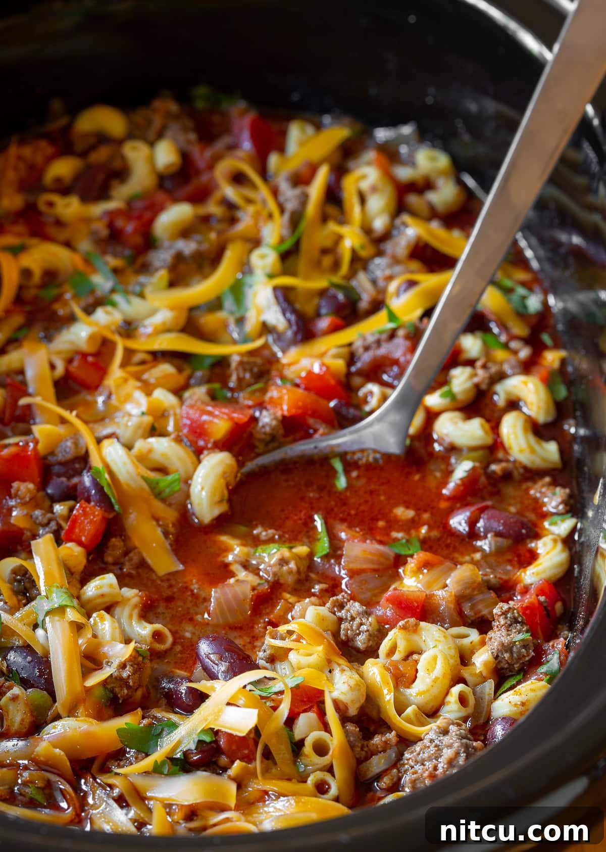 A metal ladle scoops a generous portion of hearty Slow Cooker Chili Mac and Cheese from a slow cooker, showcasing the rich, cheesy, and meaty texture.