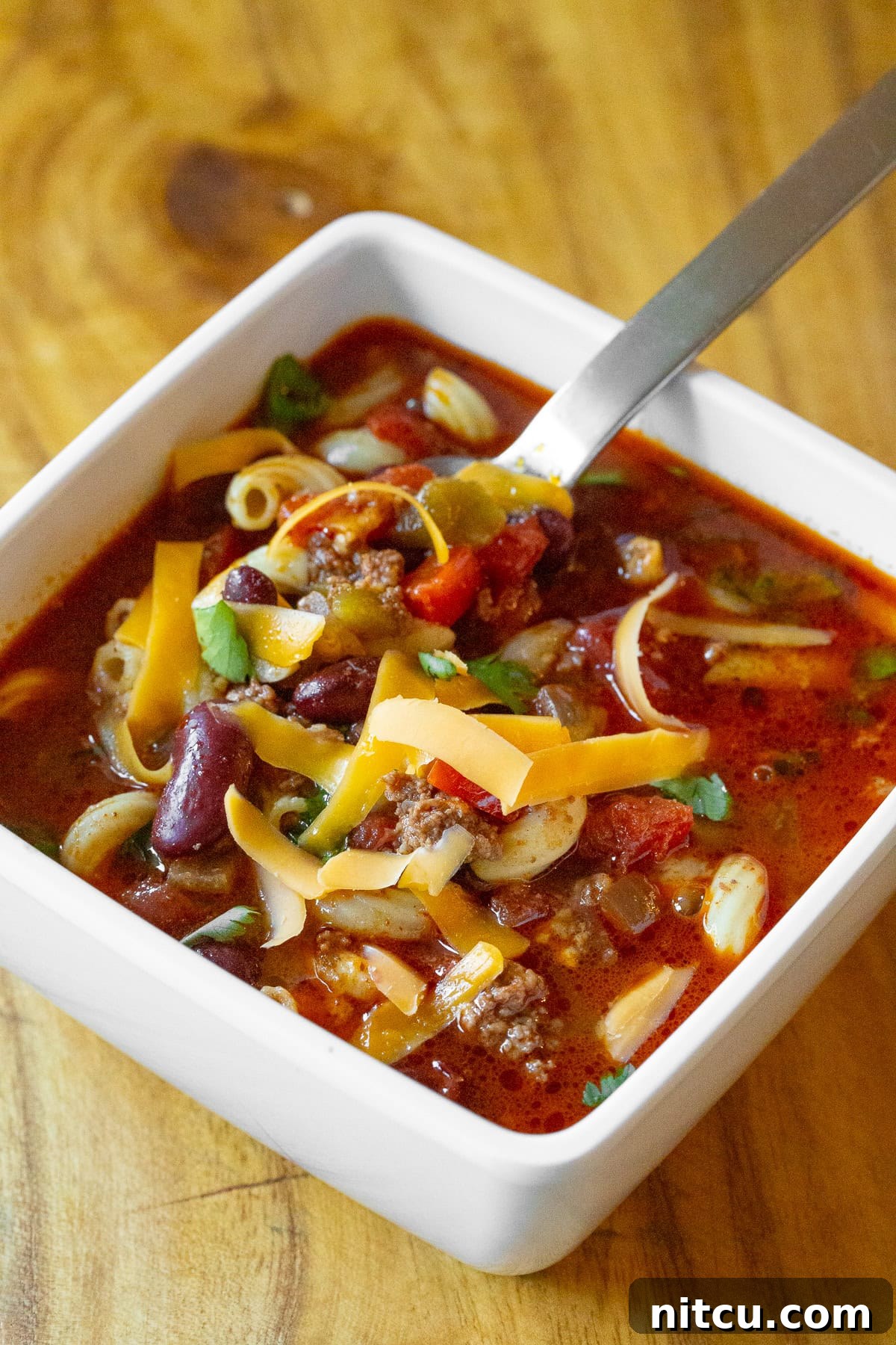 A top-down view of a single serving of Slow Cooker Chili Mac and Cheese in a bowl, with a spoon nestled in, inviting you to dig in and enjoy the hearty, cheesy goodness.