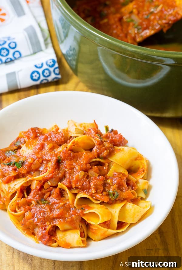 A white plate showcasing pappardelle pasta generously topped with rich, fresh homemade tomato sauce and a sprig of basil.