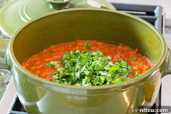 A hand adding fresh chopped basil into simmering tomato sauce in a vibrant green pot, enhancing its aroma and flavor.