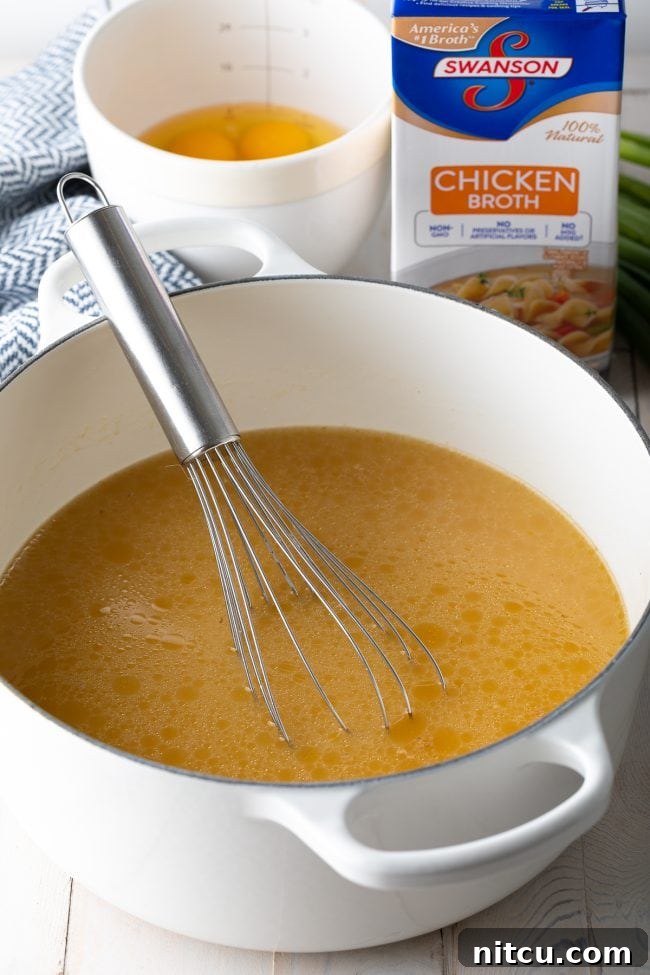 Cozy Kitchen Egg Drop Soup 4 Pot of broth in a large white cooking pot, with a metal whisk in the pot. In the background are a box of chicken broth and a white mixing bowl with eggs.