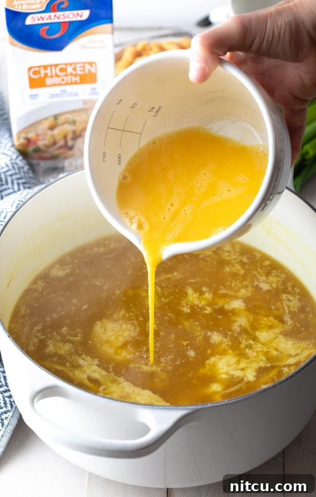 Cozy Kitchen Egg Drop Soup 5 Adding whisked eggs from a white measuring bowl into a pot of broth.