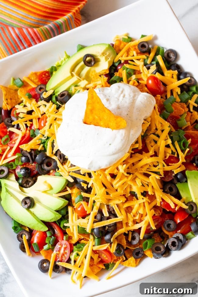 Legendary Dorito Taco Salad 2 A vibrant Dorito taco salad, richly loaded with crisp lettuce, halved grape tomatoes, thinly sliced black olives, chopped green onions, creamy avocado slices, and generous shredded cheddar cheese, all topped with a dollop of cool ranch sour cream and a signature Dorito chip.