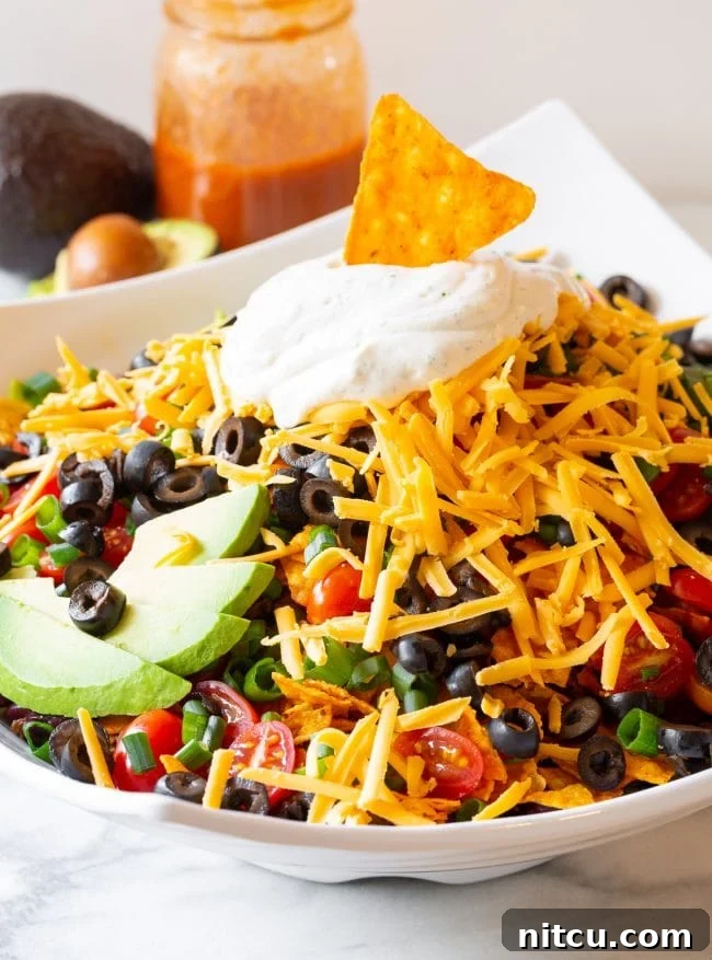 Legendary Dorito Taco Salad 11 A close-up of a generously loaded Dorito taco salad bowl, showcasing crisp lettuce, vibrant grape tomatoes, sliced black olives, green onions, avocado, shredded cheddar, and dollops of ranch sour cream, with a Dorito chip serving as a garnish.