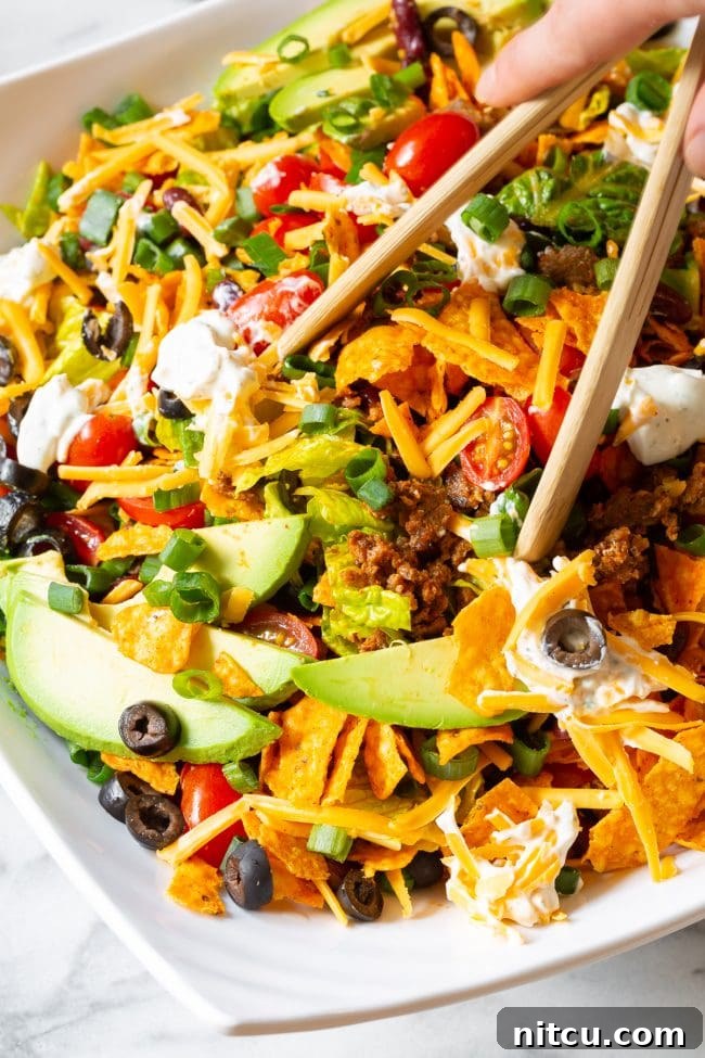 Legendary Dorito Taco Salad 4 A hand using wooden tongs to gently toss a large Dorito taco salad, revealing layers of crushed Dorito chips and fresh salad fixings.