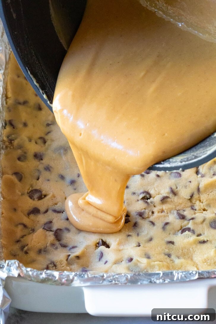 Caramel sauce being poured onto a layer of cookie dough in a baking dish.
