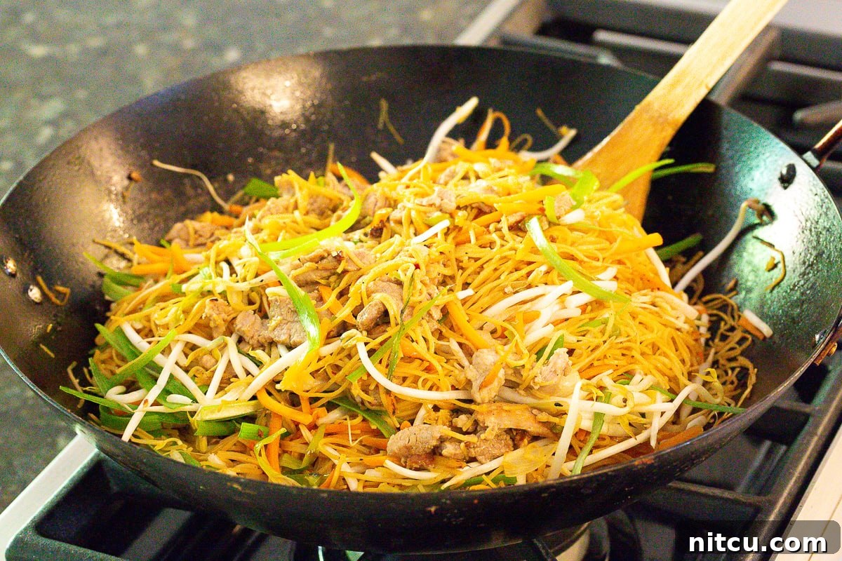 Savory Cantonese Pork Noodles 7 A chef expertly stir-frying noodles, pork, and vegetables in a large wok, creating a delicious pan-fried noodle dish.