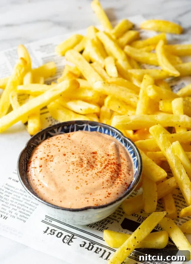 The Encore Sauce 5 A vibrant top-down view of Southern Comeback Sauce in a bowl, flanked by golden French fries and laid out on a sheet of newspaper for a rustic feel.