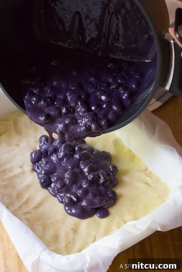 Rich, purple blueberry sauce being poured from a pot onto the cookie base in a parchment-lined baking dish.