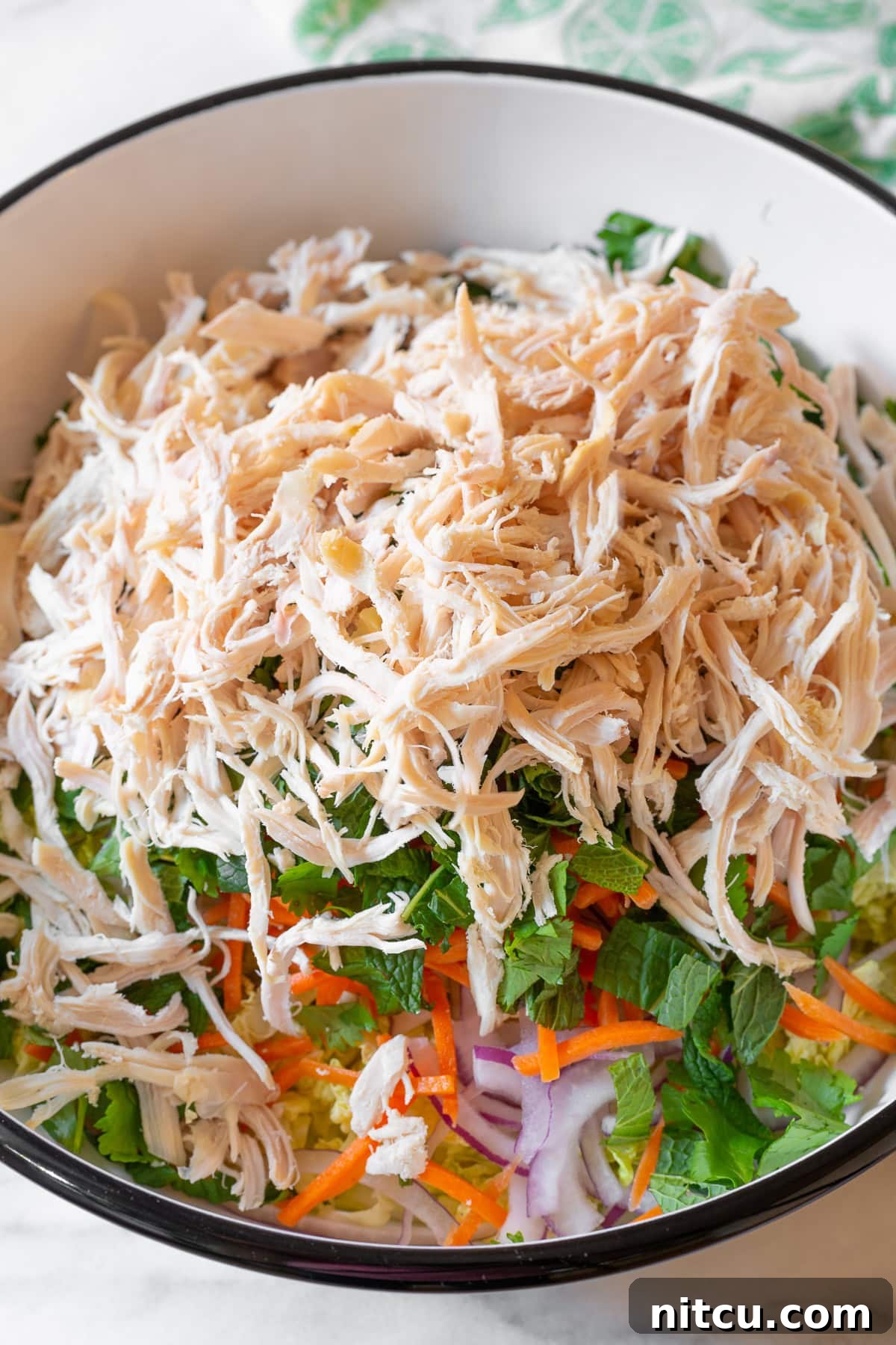 Freshly shredded chicken breast elegantly layered on top of a colorful mix of crisp vegetables in a large mixing bowl, awaiting the final dressing for Goi Ga.