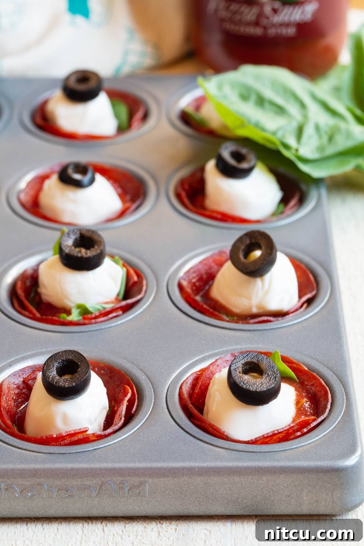 Savory Pepperoni Crisps 5 A mini muffin pan showcases pepperoni cups, each meticulously filled with fresh basil, zesty pizza sauce, a mozzarella ball, and sliced olives, eagerly awaiting their final bake.
