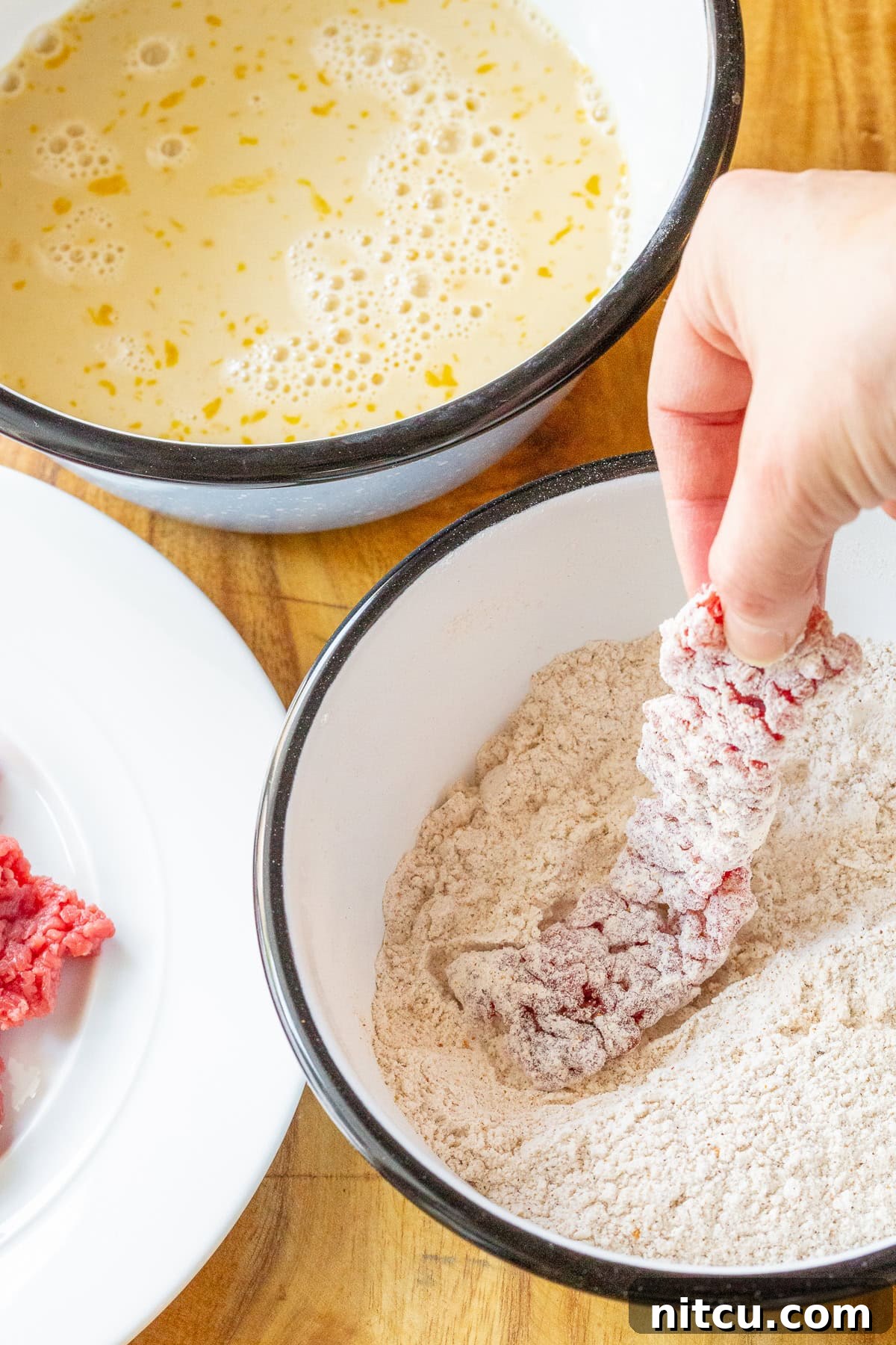 Beef Tenders 4 A hand dipping a strip of raw beef into a bowl of batter and then into a bowl of seasoned flour, preparing it for frying.