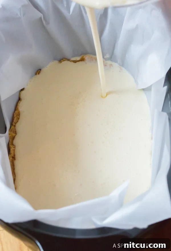 A close-up of cheesecake ingredients being poured into a slow cooker liner, highlighting the smooth texture of the filling over the crust.