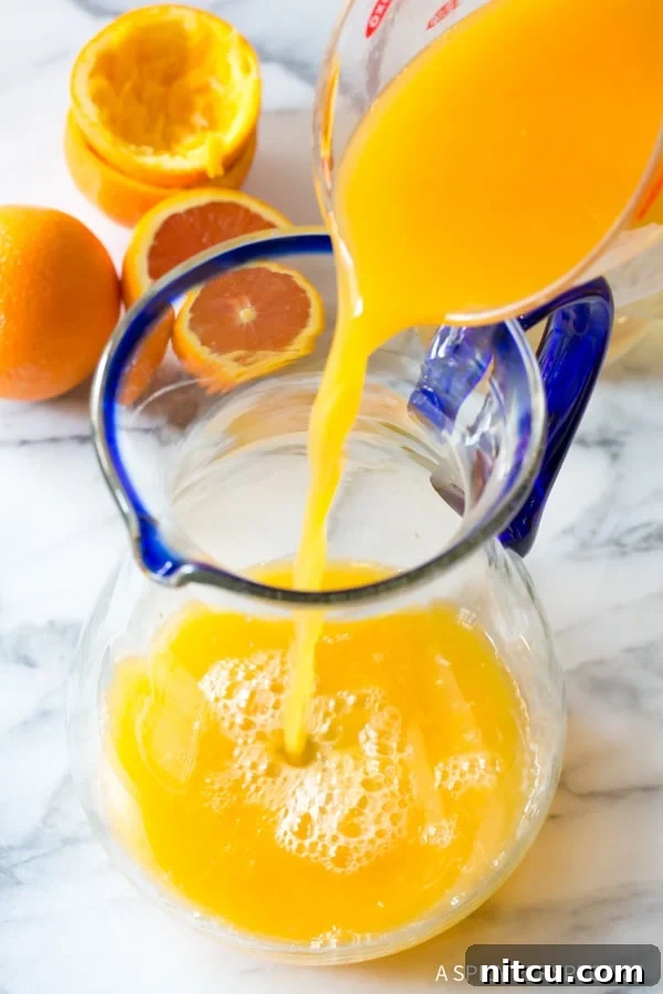 Adding Fresh Orange Juice to Orangeade Mixture Freshly squeezed orange juice, a rich golden hue, being carefully poured into a large clear glass pitcher, symbolizing the start of a homemade orangeade batch.
