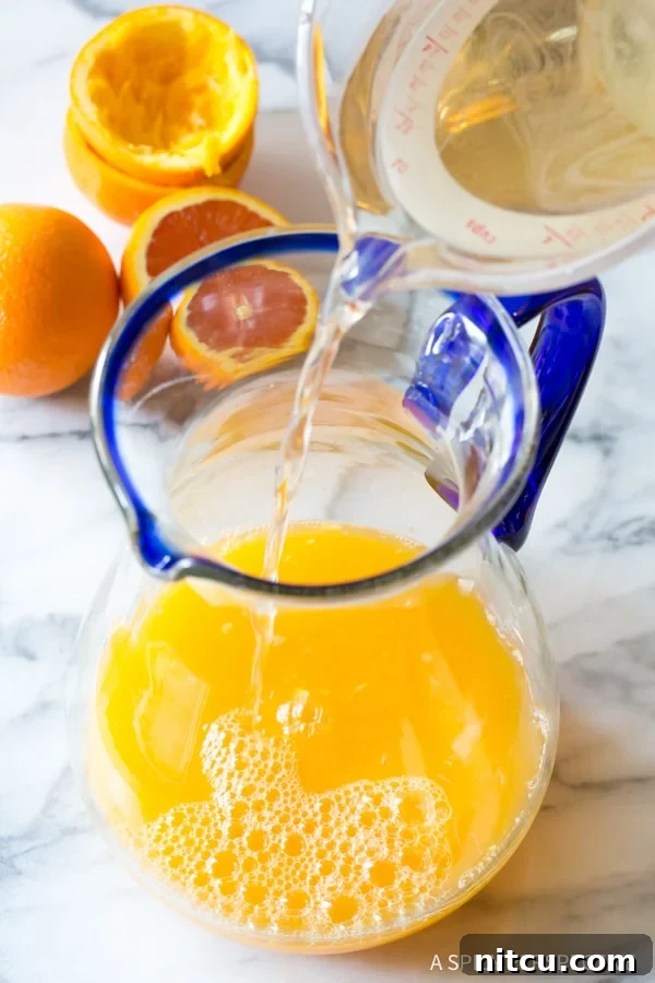 Mixing Simple Syrup for Orangeade - Sweetener Step A stream of clear simple syrup being poured into a large glass pitcher containing citrus juices, an essential step in preparing homemade orangeade.