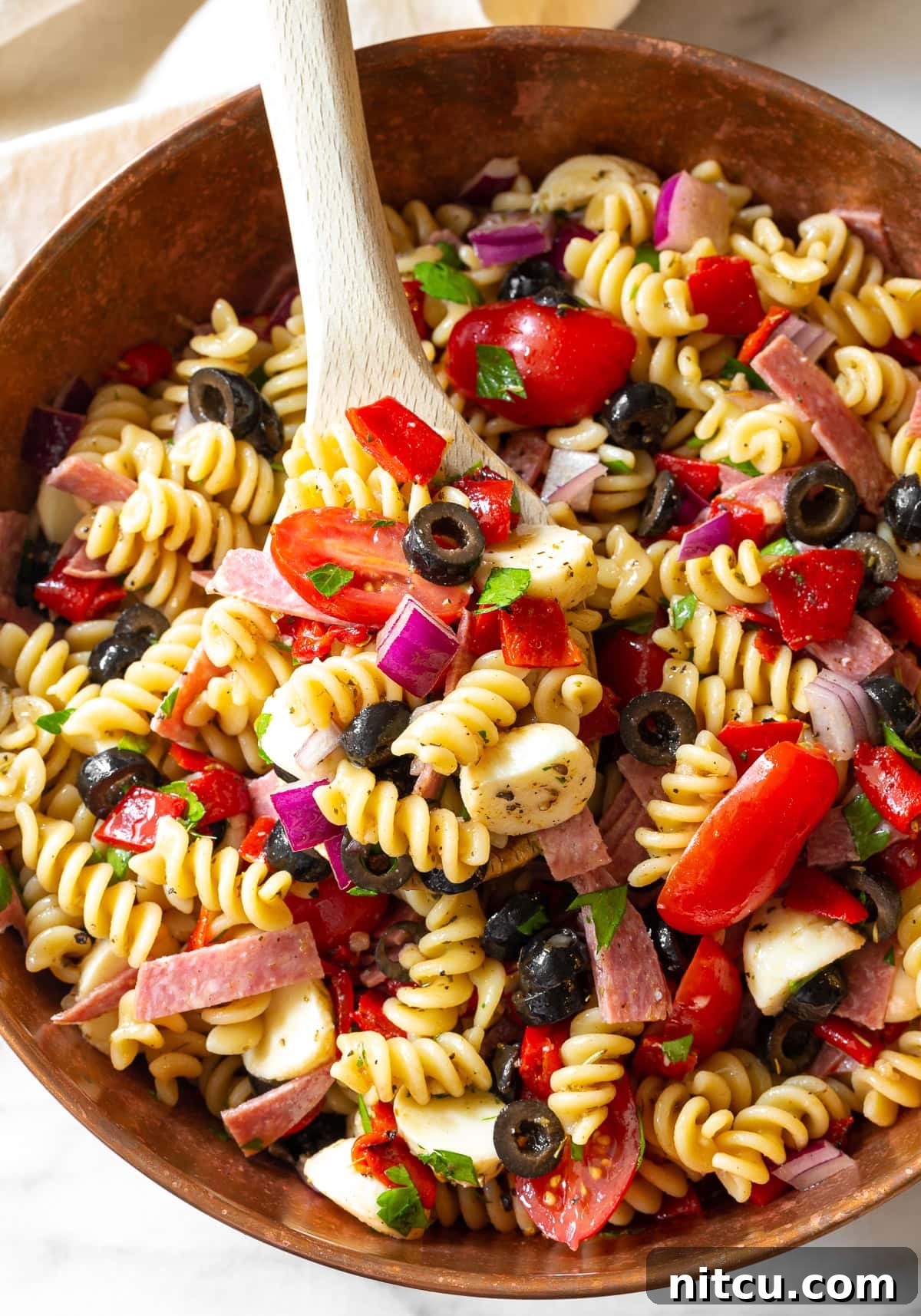 A large bowl of vibrant Italian pasta salad, filled with colorful vegetables, salami, and mozzarella, with a wooden spoon ready for serving.