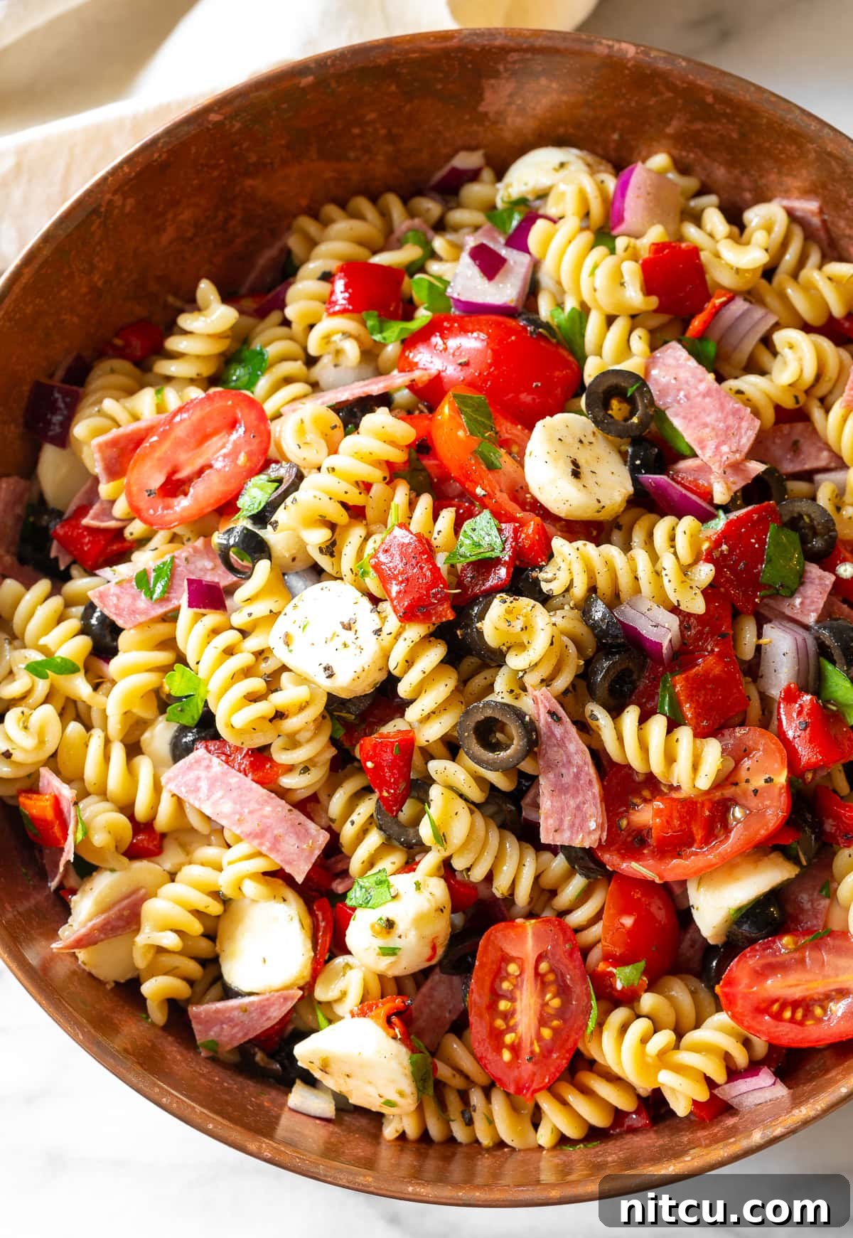 Large bowl of Italian pasta salad, featuring a rich mix of pasta, veggies, salami, cheese, and olives, glistening with dressing.