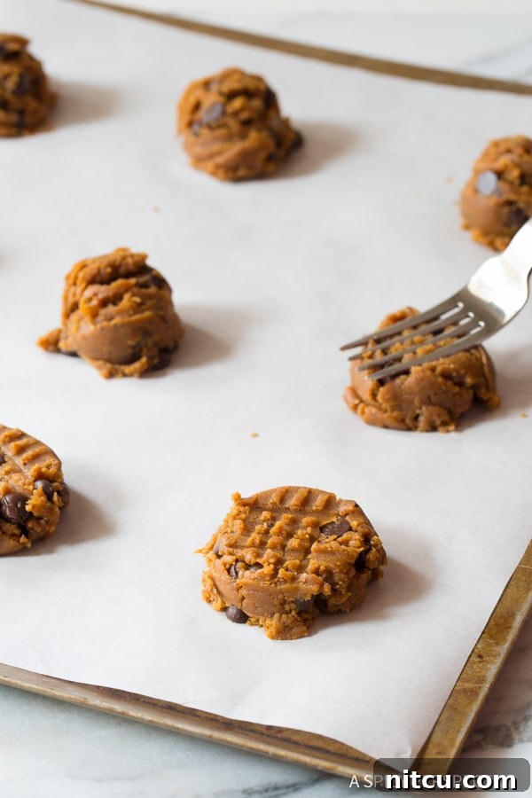 Magic 4-Ingredient Guilt-Free Chocolate Chip Cookies 7 A baker using a fork to gently press down on individual cookie dough balls on a baking sheet, creating the classic crosshatch pattern.