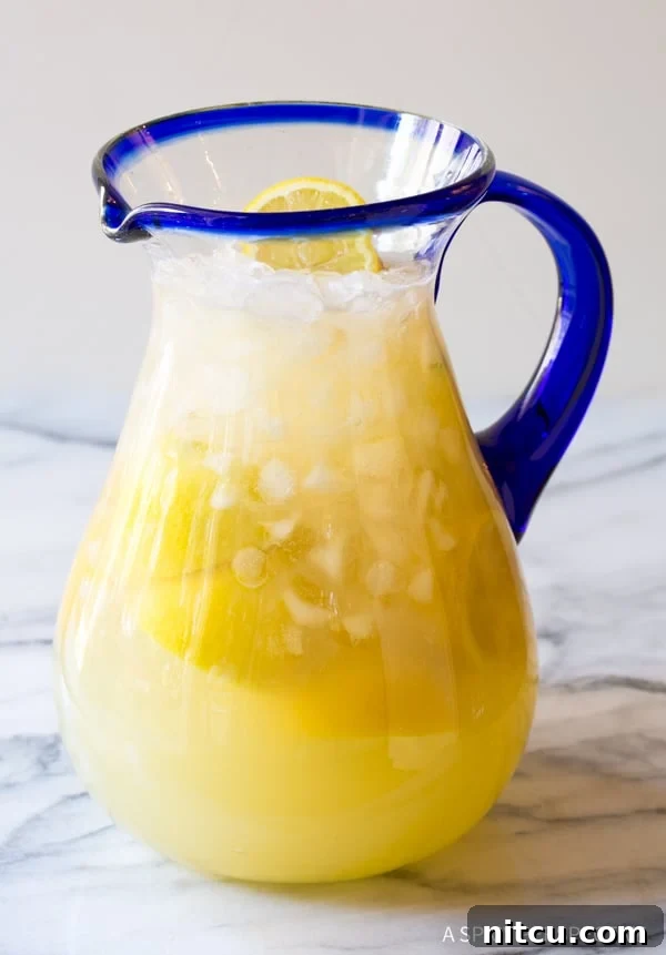 Naturally Sweet Lemonade 5 Side view of a clear glass pitcher brimming with vibrant Stevia Lemonade, artfully garnished with fresh lemon slices and a sprig of mint, resting on a rustic wooden surface.