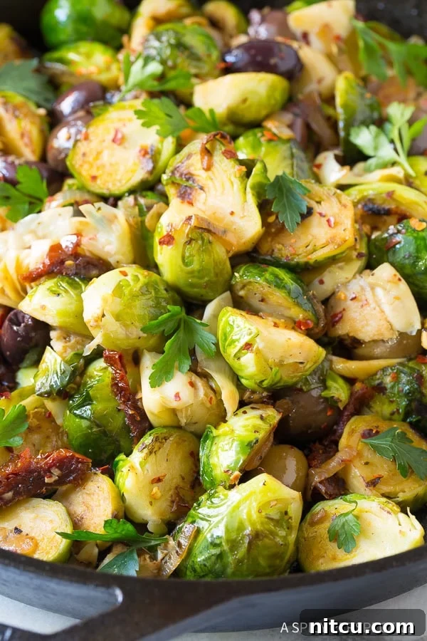 Close view of braised brussels sprouts with sun-dried tomatoes, greek olives, artichoke hearts and fresh chopped parsley, glistening in a skillet.
