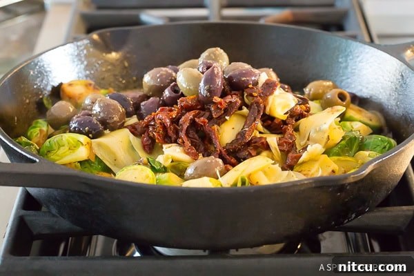 Adding sun-dried tomatoes, greek olives, and artichoke hearts to the cast iron skillet with brussel sprouts, mid-cooking.