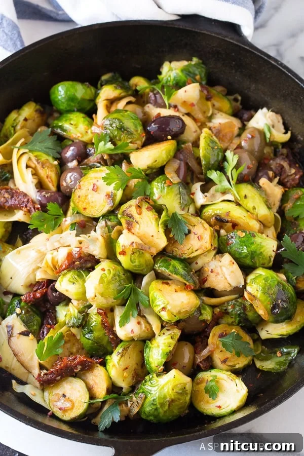 Braised brussels sprouts with sun-dried tomatoes, greek olives, artichoke hearts and fresh chopped parsley in a cast iron skillet, garnished and ready to serve.