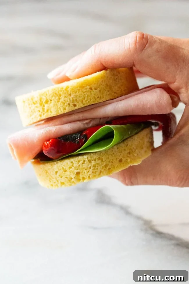 Magic Mug Low Carb Bread Recipe #ASpicyPerspective #Paleo #Ketogenic #SugarFree #DairyFree #GrainFree #GlutenFree Hand holding a small sandwich made with two slices of low carb bread, sandwiching deli meat, roasted red pepper, and spinach leaves.